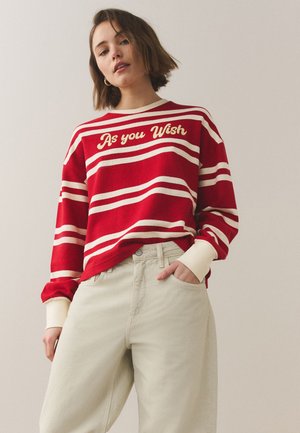 Ung kvinde iført rød og hvid striped sweater med teksten "As you Wish", kombineret med højtaljede lyse beige bukser, stående mod en ensfarvet baggrund.