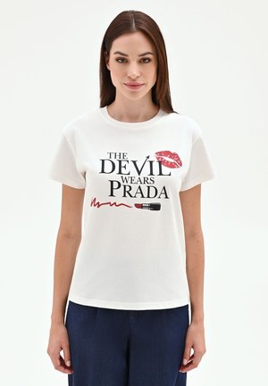 Femme aux cheveux bruns et raides portant un T-shirt blanc avec "The Devil Wears Prada", une marque de rouge à lèvres rouge et un motif de rouge à lèvres, pantalon foncé.
