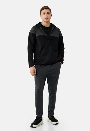 Młody mężczyzna stoi, ubrany w czarną bluzę z kapturem na suwak z szarymi panelami, ciemnoszare spodnie oraz czarno-białe sneakersy na jednolitym tle.