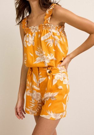 Woman wearing orange sleeveless ruffled top and matching floral shorts with buttoned belt detail, posing with one hand on hip.