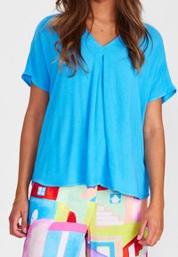 Blusa leggera azzurro chiaro a maniche corte con scollatura a V e piega centrale. Tessuto liscio e vestibilità comoda, abbinata a pantaloni colorati a motivi.