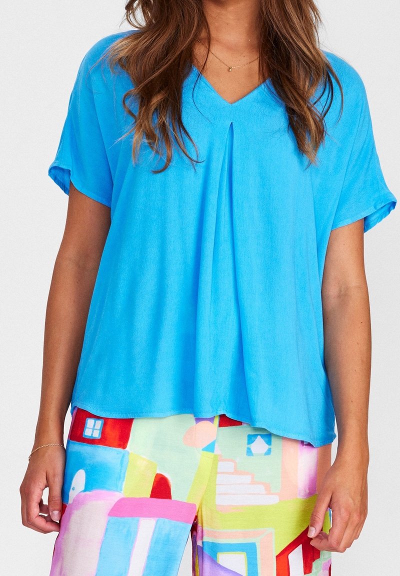 Blusa leggera azzurro chiaro a maniche corte con scollatura a V e piega centrale. Tessuto liscio e vestibilità comoda, abbinata a pantaloni colorati a motivi.