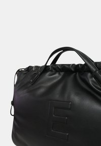 Borsa in pelle nera con parte superiore arriccita, caratterizzata da una grande lettera "E" in rilievo sul fronte. Texture liscia e manici strutturati.