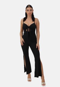 Combinaison noire côtelée avec un devant noué, un décolleté en V profond et des jambes évasées avec des fentes sur les côtés. Associée à des talons blancs à lanières.