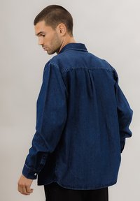 Mørkeblå denimskjorte med avslappet passform, lange ermer og mansjetter med knapper. Har et bakstykke og en subtil tekstur.