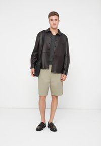 Sötétbarna bőr ingdzseki, szürke pulóver, bézs rövidnadrág és fekete slip-on cipő. A modell egy fekete válltáskát tart.