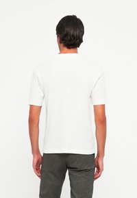 Man with dark hair wearing a plain white short-sleeve shirt and dark gray pants, standing facing a white background.