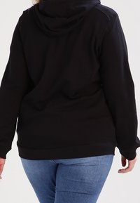 Felpa nera con finitura testurizzata, cappuccio attaccato e orlo a costine. Indossata con denim blu, che mostra una vestibilità rilassata e un design casual.
