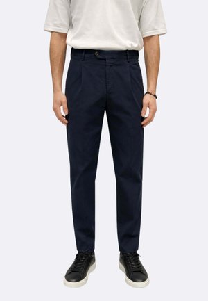 Uomo con pantaloni sartoriali blu scuro, camicia bianca a maniche corte, scarpe da tennis nere con suole bianche e braccialetto nero con perline in piedi.