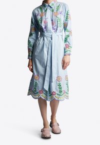 Abito camicia azzurro chiaro con ricami floreali in rosa, viola e verde. Presenta una vita legata e un orlo scallop con tagli.