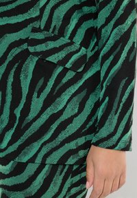 Tessuto a righe verde e nero con una finitura testurizzata, caratterizzato da una tasca su un indumento su misura, evidenziando un design moderno a stampa animale.