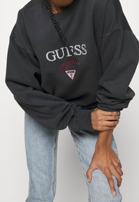 Sudadera de color gris carbón con el texto "GUESS" y el logotipo en bordado rojo y plateado. Presenta un corte relajado y puños acanalados. Combinada con jeans de color azul claro.