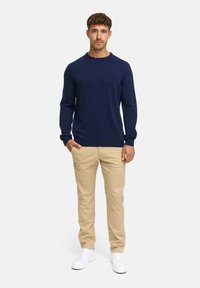 Marineblå strikket genser med rund hals og lange ermer, kombinert med lyse khaki chinos og hvite joggesko, stående mot en hvit bakgrunn.