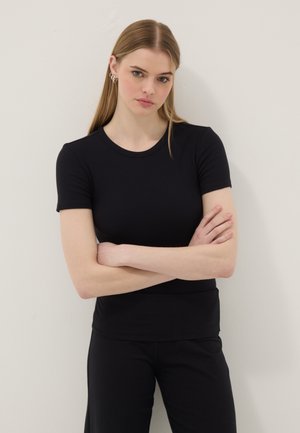 Junge Frau mit glatten blonden Haaren und heller Haut, trägt ein eng anliegendes schwarzes Kurzarmoberteil und schwarze Hosen, Arme verschränkt, neutraler Hintergrund.