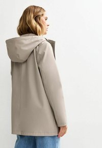 Veste imperméable beige avec capuche, coupe droite et détails de couture visibles, associée à un jean en denim bleu. La vue arrière montre une texture lisse.