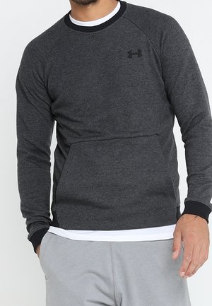 Man wearing dark gray Under Armour long-sleeve sweatshirt over white shirt, with light gray pants, hand in pocket.