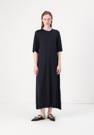 Schwarzes knielanges Kleid aus glattem Stoff mit kurzen Ärmeln und rundem Ausschnitt, kombiniert mit schwarzen flachen Schuhen mit Schleifen.