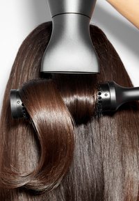 Outil de coiffure noir et argent avec un cylindre arrondi, mèches de cheveux brillants et châtain, mettant en valeur une texture lisse et des boucles élégantes.