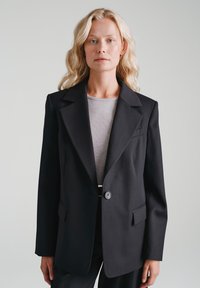 Czarna, dopasowana marynarka o strukturalnym kroju, z klapami z wycięciem, pojedynczym zapięciem na guzik i dwoma przednimi kieszeniami. Noszona na szarym topie.