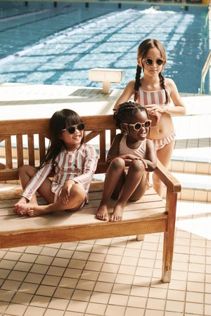Τρία παιδιά με ριγέ μαγιό κάθονται σε ένα ξύλινο παγκάκι δίπλα σε μια πισίνα. Απαλοί χρωματισμοί, γυαλιά ηλίου, νερό στο φόντο και πλακάκια στο πάτωμα.