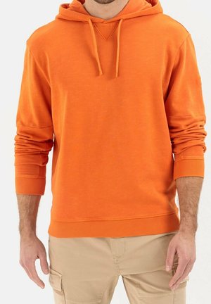 Man draagt een feloranje hoodie met trekkoorden en beige cargobroek, staand tegen een effen lichte achtergrond.