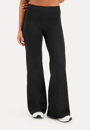 Pantaloni neri a gamba larga indossati con sneakers bianche, visibili dalla vita ai piedi su uno sfondo chiaro e uniforme.