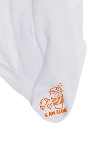 Weißes Kleidungsstück aus Stoff mit einer orangefarbenen Grafik von einer Kaffeetasse, einem Croissant und dem Text "6 AM CLUB" in der Nähe des Saums.