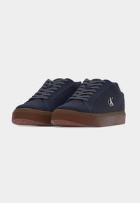 Paire de baskets basses bleu marine avec semelles en caoutchouc marron et lacets gris, avec un logo blanc "CK" sur le côté.