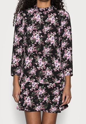 Zwarte jurk met roze en paarse bloemenprint, lange mouwen en een gerimpelde zoom, gedragen door een staand persoon met krullend haar.