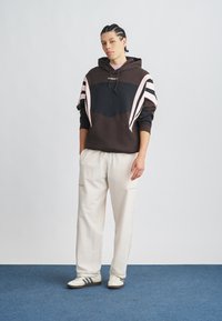 Fiatal személy áll egy kék szőnyegen, fekete és rózsaszín Adidas kapucnis pulóvert, világos bézs nadrágot és fehér, fekete csíkos tornacipőt viselve.