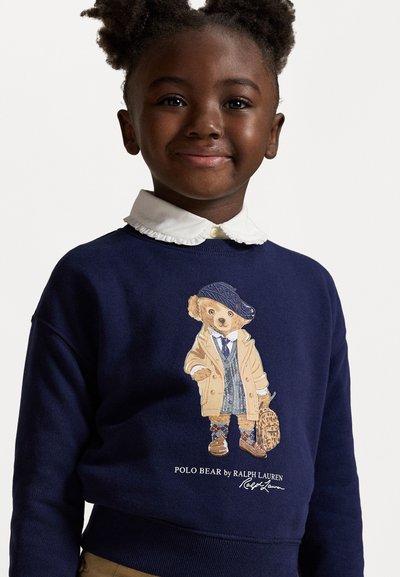 Marinfärgad sweatshirt med en grafik av en björn i en beige kappa, blå rutig väst och keps, accentuerad av en vit skjorta under.