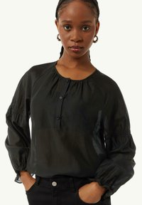 Schwarze Bluse mit Knopfleiste, rundem Ausschnitt, Ballonärmeln und gerafften Details, aus leichtem Stoff. Getragen mit schwarzen Hosen.