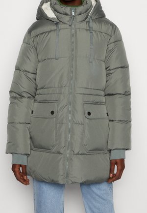 Mand iført grå quiltet vinterjakke med hætte og frontlynlås, to klaplommer og lyseblå jeans, stående mod en hvid baggrund.