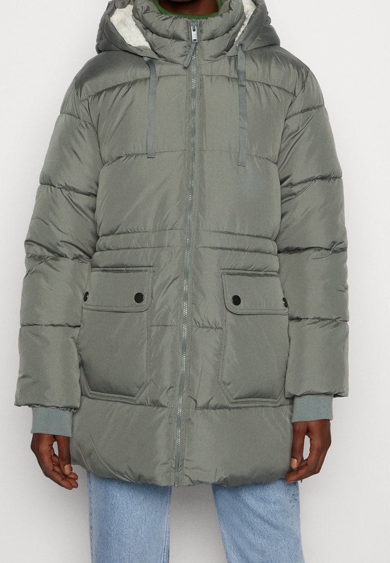 Homme portant une veste d'hiver matelassée grise à capuche avec une fermeture éclair à l'avant, deux poches à rabat, et un jean bleu clair, debout devant un fond blanc.