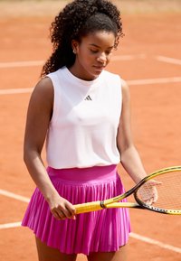 Top blanco sin mangas con textura y escote redondo, combinado con una falda rosa plisada. Sosteniendo una raqueta de tenis amarilla en una cancha de tierra batida.