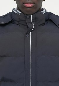 Marineblauer gepolsterter Jacke mit Stehkragen, weißen Akzenten und Knopfverschluss. Glatte Textur mit sichtbaren Steppdetails.