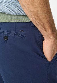 Marineblauwe herenbroek van een gladde, lichte stof. Voorzien van een enkele zwarte knop en een logo-label op de achterzak.