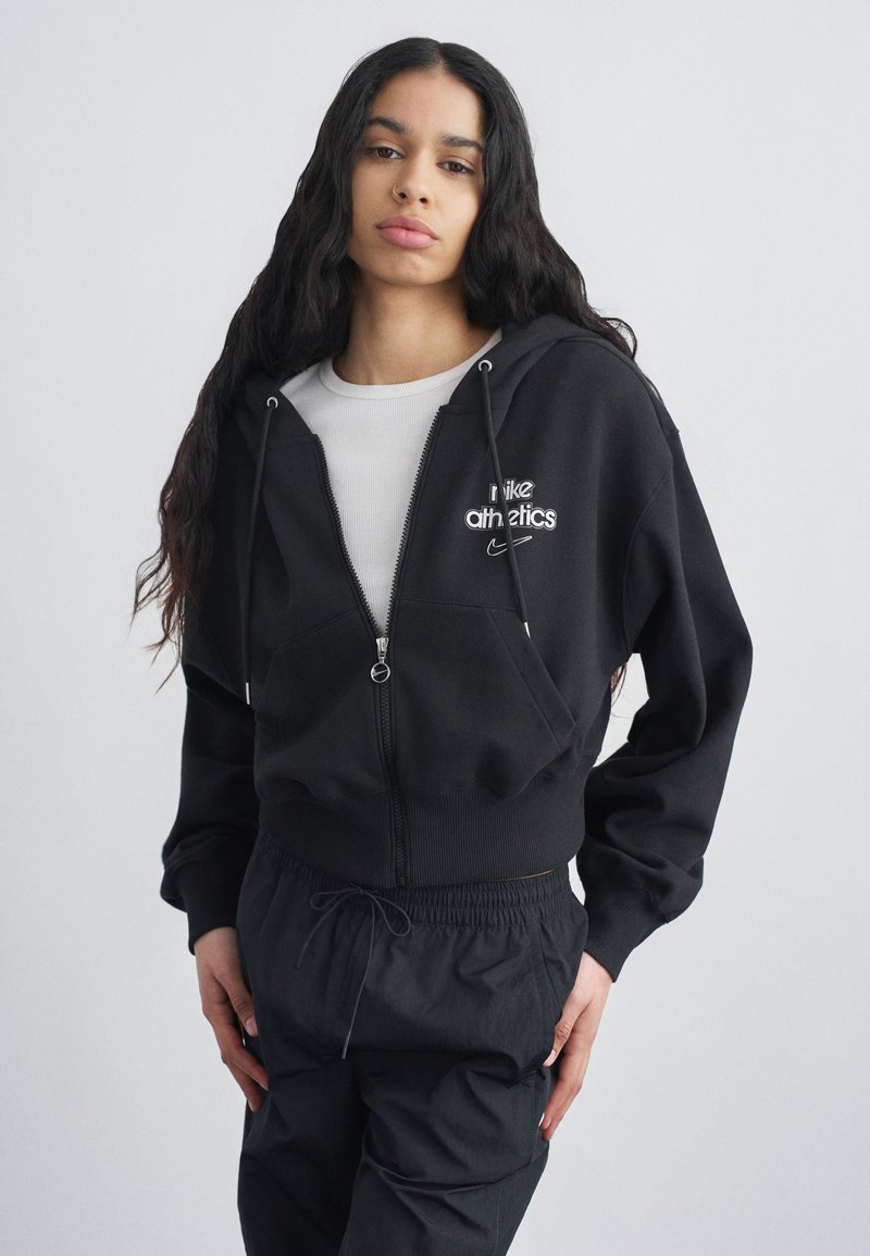 Νεαρή γυναίκα με μακριά σκούρα μαλλιά, ντυμένη με μαύρο φούτερ Nike Athletics με φερμουάρ και μαύρο παντελόνι φόρμας, μπροστά σε απλό φόντο.