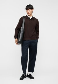 Καφέ πουλόβερ πλεκτό με γιακά, συνδυασμένο με σκούρο μπλε παντελόνι με στενή γραμμή. Το άτομο κρατά μια μαύρη δερμάτινη τσάντα tote και φοράει μαύρα loafers.