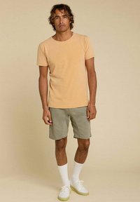 T-shirt à col rond orange clair, shorts verts olive, chaussettes blanches et baskets blanches. Design simple avec des accents minimaux.