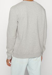 Man in een lichtgrijze longsleeve sweatshirt en lichtblauwe jeans, gezien vanuit de rug tegen een effen achtergrond.