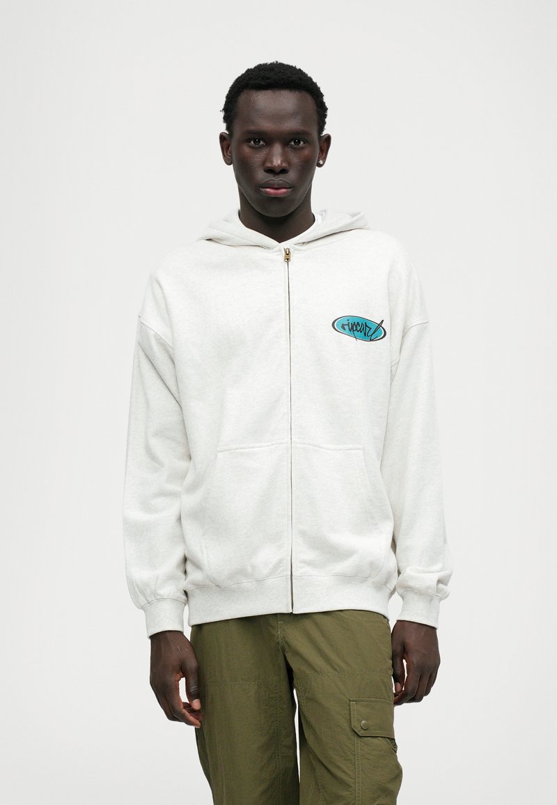 Helehall hallikleebiga kapuutsiga jakk, millel on ees taskud, soonikkoes varrukad ja teal värvi logo. Kantakse roheliste cargo pükste peal. Sile tekstuur, lihtne lõige.