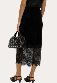 Zwarte handtas met zilveren studs, van gestructureerd leer. Model draagt een zwarte velours rok met kanten rand en zwarte hoge hakken.