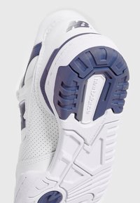 Vit träning shoe med perforerat läderöverdel, blå accenter. Har en texturerad gummisula med greppmönster och varumärke på undersidan.