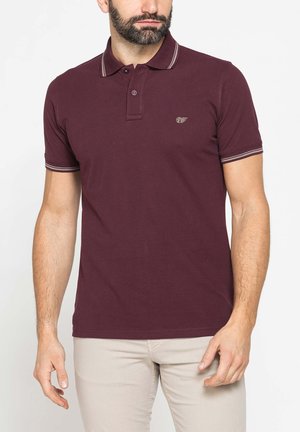 Uomo che indossa una polo a maniche corte rosso scuro con collo e polsini a righe, abbinata a pantaloni beige chiaro su uno sfondo semplice.