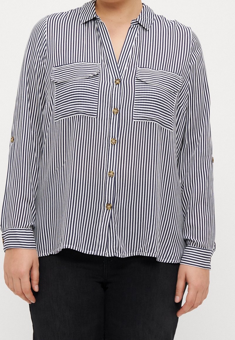 Personne portant une chemise à rayures verticales noires et blanches boutonnée, avec manches longues et deux poches poitrine sur un pantalon noir.