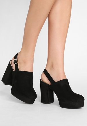 Zwarte suède platform klompen met een dikke blokhak en open achterkant. Voorzien van een verstelbare band met een metalen sluiting als accent.