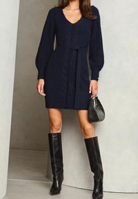 Mörkblå stickad klänning med V-ringning, kabeldetaljer, puffärmar, knytning i midjan och svarta knähöga stövlar. Tillbehör inkluderar en svart väska.