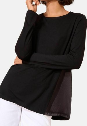 Kobieta ubrana w czarną bluzkę z długim rękawem z przezroczystymi panelami po bokach oraz białe spodnie, pozująca z jedną ręką skrzyżowaną na klatce piersiowej, a drugą dłonią przy twarzy.