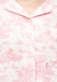 Chemise blanche avec un motif toile de Jouy rose présentant des scènes pastorales détaillées et quatre boutons blancs sur le devant.